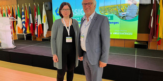 Katharina Schrot und Jürgen Schultze von der Sozialforschungsstelle vertreten Evolving Regions auf dem "Forum on Mission Adaptation to Climate Change". Im Hintergrund hinter ihnen sind Flaggen der EU-Staaten aufgestellt. 