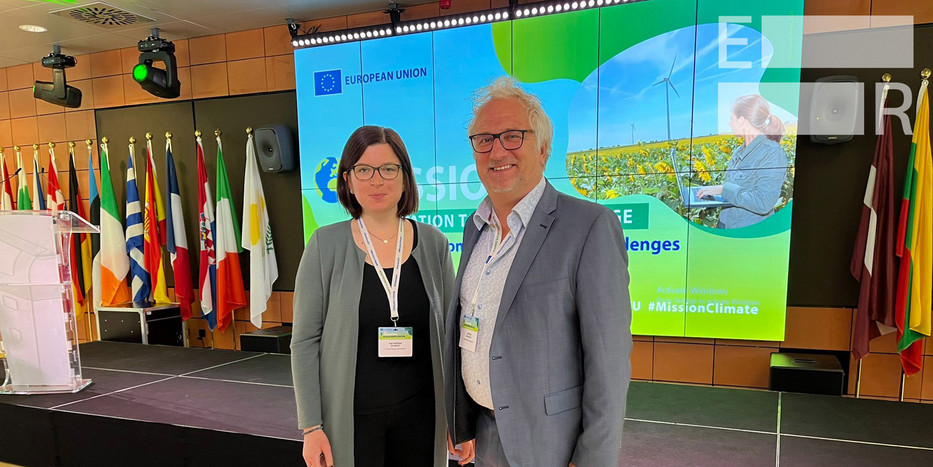 Katharina Schrot und Jürgen Schultze von der Sozialforschungsstelle vertreten Evolving Regions auf dem "Forum on Mission Adaptation to Climate Change". Im Hintergrund hinter ihnen sind Flaggen der EU-Staaten aufgestellt. 