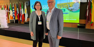 Katharina Schrot und Jürgen Schultze von der Sozialforschungsstelle vertreten Evolving Regions auf dem "Forum on Mission Adaptation to Climate Change". Im Hintergrund hinter ihnen sind Flaggen der EU-Staaten aufgestellt.