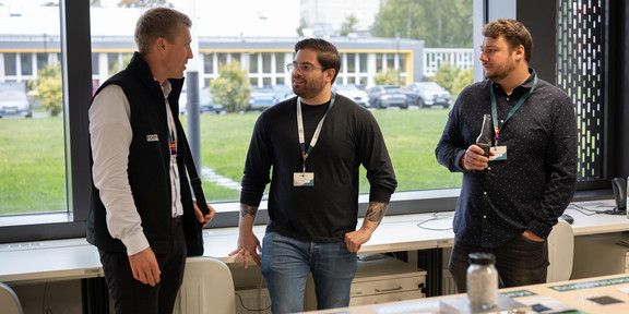Antonio Isopp steht vor einem Tisch, auf dem Dokumente liegen. Er unterhält sich mit zwei weiteren Personen, die links und rechts von ihm stehen.