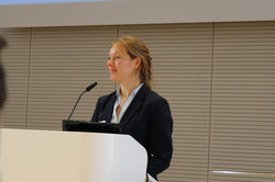 Eine Person steht an einem Rednerpult in einem Konferenzraum und spricht vor Publikum. Auf der Leinwand über ihr ist ein Präsentationsausschnitt zu sehen.