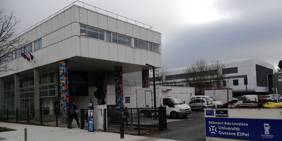 Foto: Gebäude mit der Beschriftung „Bâtiment Ada Lovelace – Université Gustave Eiffel“. Vor dem Eingang stehen Fahrzeuge und Personen, daneben ein Schild mit weiteren Informationen zur Universität.