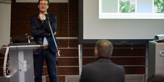 Bastian Pelka steht neben einem Pult vor einem Mikrofon.