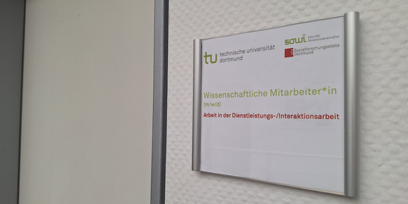 Foto: Türschild mit Logos der TU Dortmund, der Fakultät Sozialwissenschaften und der sfs. Darunter der Text "Wisseschaftliche Mitarbeiter*in (m/w/d)" und "Arbeit in der Dienstleistungs-/Interaktionsarbeit". Neben dem Türschild ist eine geschlossene Tür.
