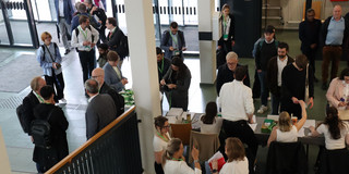 Foto: Personen stehen und unterhalten sich im Eingangsbereich einer Veranstaltung, einige sind an einem Empfangstisch beschäftigt.