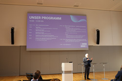 Jürgen Schultze spricht in ein Mikrofon, dass er in der Hand hält. Hinter ihm wird das Programm der Konferenz auf eine Leinwand projiziert. Vor ihm sitzt eine Zuhörerschaft.
