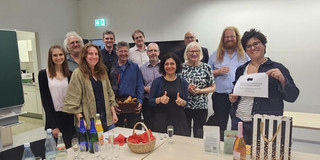 Karina is in the center of the picture. Her friends, coworkers, and family are surrounding her. In front of them is a table set with sparkling wine glasses, drinks, and gifts.
