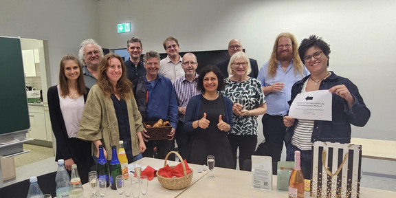Karina is in the center of the picture. Her friends, coworkers, and family are surrounding her. In front of them is a table set with sparkling wine glasses, drinks, and gifts.
