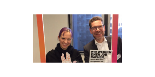 Kerstin Guhlemann und Prof. Dr. Sascha Stowasser stehen hinter einem roten Rahmen. Unten am Rahmen ist das Modell einer Hand befestigt, die mit ihren Fingern ein "V" formt. Daneben ist ein kleines Schild angebracht, auf dem "WIR WERDEN EINEN JOB MACHEN, den die Welt noch nicht gesehen hat." steht.
