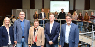 Foto: Gruppe von etwa 15 Personen auf zwei Ebenen an einer runden Innenbrüstung vor einer Projektionsleinwand; fünf Personen stehen vorne an der Brüstung, die übrigen auf der Galerie.