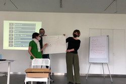 Foto: Personen präsentieren ein Plakat in einem Seminarraum mit Flipchart und Projektionsfläche.