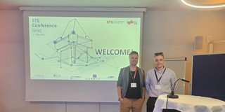 Two people are standing next to a screen onto which a presentation is being projected.