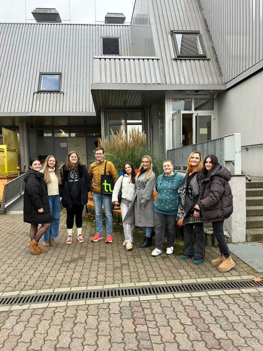 Foto: Eine Gruppe von Personen steht vor einem Gebäude mit metallverkleideter Fassade und Eingangsbereich.