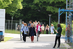 Foto: Gruppe von Personen geht auf einem gepflasterten Campusweg zwischen Gebäuden und Bäumen auf die Kamera zu.