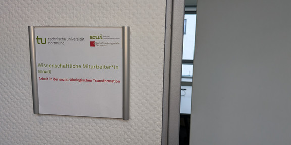 Stellenanzeige WiMi 21-26 Foto: Türschild mit Logos der TU Dortmund, der Fakultät Sozialwissenschaften und der sfs. Darunter der Text "Wisseschaftliche Mitarbeiter*in (m/w/d)" und "Arbeit in der sozial-ökologischen Transformation". Neben dem Türschild ist eine Tür einen Spalt weit geöffnet.