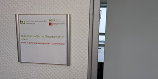 Foto: Türschild mit Logos der TU Dortmund, der Fakultät Sozialwissenschaften und der sfs. Darunter der Text "Wisseschaftliche Mitarbeiter*in (m/w/d)" und "Arbeit in der sozial-ökologischen Transformation". Neben dem Türschild ist eine Tür einen Spalt weit geöffnet.