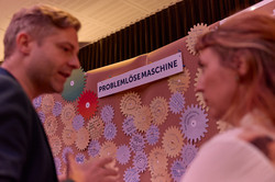 Zwei Personen stehen vor einer Pinnwand und diskutieren miteinander. Auf der Pinnwand hängt oben mittig ein Zettel mit der Aufschrift "Problemlösemaschine". Auf dem Rest der Pinnwand sind Zettel in Form von Zahnrädern befestigt. Sie sind mit Wörtern und Zeichnungen beschriftet.