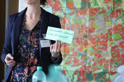 Foto: Person hält eine beschriftete Karte vor einer großformatigen Stadt- oder Plankarte; Namensschild am Revers und Wasserflasche auf dem Tisch.