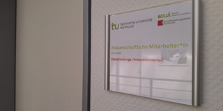 Foto: Türschild mit Logos der TU Dortmund, der Fakultät Sozialwissenschaften und der sfs. Darunter der Text "Wisseschaftliche Mitarbeiter*in (m/w/d)" und "Dienstleistungs-/Interaktionsarbeit". Neben dem Türschild ist eine geschlossene Tür.