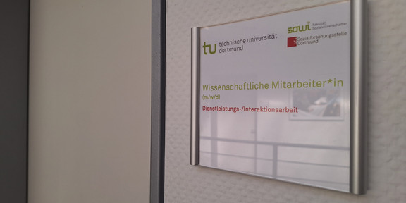 Stellenausschreibung WiMi w40/26 Deutsch Foto: Türschild mit Logos der TU Dortmund, der Fakultät Sozialwissenschaften und der sfs. Darunter der Text "Wisseschaftliche Mitarbeiter*in (m/w/d)" und "Dienstleistungs-/Interaktionsarbeit". Neben dem Türschild ist eine geschlossene Tür.