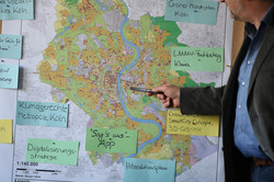 Foto: Hand mit Zeigestift zeigt auf eine großformatige Stadtkarte von Köln; um die Karte herum haften bunte Notizzettel mit Themen wie "Klimagerechte Metropole Köln", "Digitalisierungsstrategie" und "Hitzeschutzplan".