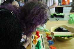Eine Person betrachtet bunte, kleine 3D-gedruckte Figuren, die auf einem Tisch ausgestellt sind. Auf dem Tisch liegen außerdem weitere Gegenstände und Materialien.