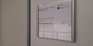 Stellenanzeige WiMi w40/26 Englisch Photo: A door sign featuring the logos of TU Dortmund University, the Faculty of Social Sciences, and the sfs. Below the logos is the text “Research Assistant (m/f/d)” and “Service/Interaction work”. Next to the door sign is a closed door.