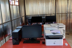 Foto. Mehrere Monitore, Computer, Drucker und Zubehör stehen auf Bierzeltgarnituren in einer Wellblechhütte.