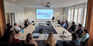 Several people are sitting at long tables in a conference room, watching a presentation projected onto the wall. One person is standing at the front speaking, while text and colored boxes are visible on the screen. Laptops, notes, and drinks are scattered across the tables.