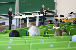Philippe Larrue steht in einem Hörsaal. Unten sind drei Stehtische aufgebaut, neben denen eine weitere Person steht. Auf den Stühlen des Hörsaals sitzen vereinzelt Personen.