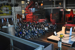 Several people sit on their chairs in the DASA steel hall and look at the empty podium.