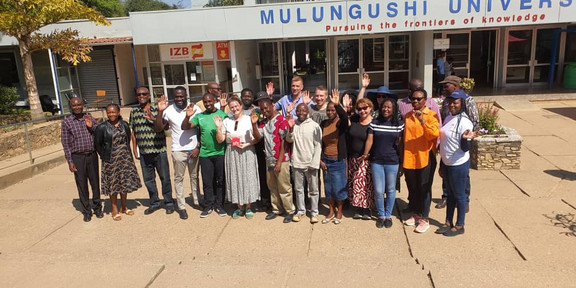 Foto: Gruppe von rund 20 Personen steht vor dem Gebäude "Mulungushi University"; viele lächeln und winken in die Kamera.