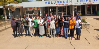 Foto: Gruppe von rund 20 Personen steht vor dem Gebäude "Mulungushi University"; viele lächeln und winken in die Kamera.