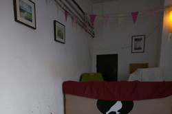 Ein kleiner Raum mit einem Bett im Hintergrund, darauf eine rote Decke und ein Kissen. Im Vordergrund steht ein beleuchtetes Schild mit der Aufschrift „The Sleeping Project – What now?“ auf einem Holzstuhl, an der Wand hängen mehrere Bilder.