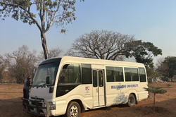 Foto: Kleinbus mit der Aufschrift "Mulungushi University" steht auf unbefestigtem Gelände unter einzelnen Bäumen; eine Person steht neben dem Fahrzeug.