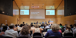 Decorative photograph of the conference plenary hall, featuring numerous people seen from behind and a screen in the distance