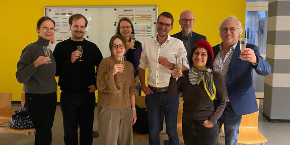 A group of people is standing in a room with a yellow wall, raising glasses for a toast.