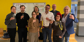 Eine Gruppe von Menschen steht in einem Raum mit gelber Wand und stößt mit Gläsern an.