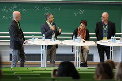 Foto: Vier Personen an Stehtischen in einem Hörsaal vor einer Tafel; eine Person spricht ins Mikrofon, Wasserflaschen auf den Tischen.
