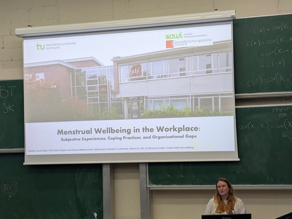 Foto: Christine Best steht in einem Hörsaal. Auf eine Leinwand wird ihre Präsentation projizziert.