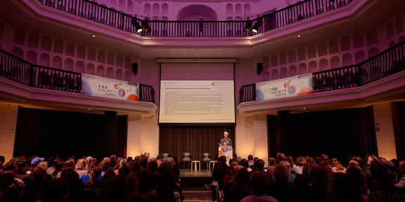 Zukunftsfabrik Bild 2 Ein Redner hält steht vor einem Podium und spricht zu einer großen Zuhörerschaft, die in einem Theater sitzt.