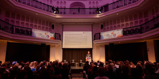 Zukunftsfabrik Image 2 A speaker stands in front of a podium and speaks to a large audience sitting in a theater.