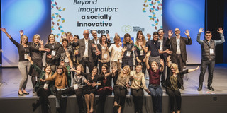 Viele Mitglieder der Social Innovation Community sitzen auf und stehen auf einer Bühne und winken lachend Richtung Kamera.