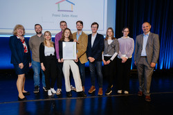 Foto: Neun Personen stehen auf einer Bühne. Zwei Personen halten eine gerahmte Urkunde in der Hand.