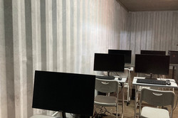 Foto. Mehrere kleine Tische stehen in mehreren Reihen nebeneinander in einem Container. Auf ihnen stehen Monitore, Tastaturen und Computermäuse.