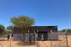 Foto einer umzäunten Hütte in einem afrikanischen Dorf. Vor und neben ihr stehen einige Personen und sehen sich die Gegebenheiten an.