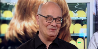 Foto: Klaus Kock steht im Studio der Fernsehsendung „Lokalzeit Dortmund“ des WDR, im Hintergrund ist ein Bildschirm mit einem Bild aus dem Alltag zu sehen.