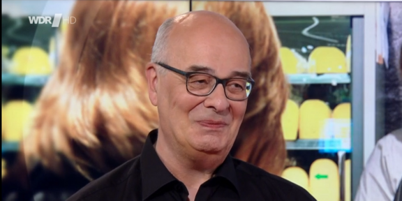 Foto: Klaus Kock steht im Studio der Fernsehsendung „Lokalzeit Dortmund“ des WDR, im Hintergrund ist ein Bildschirm mit einem Bild aus dem Alltag zu sehen.