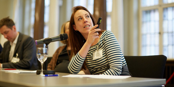 Foto: Marthe Zirngiebl spricht bei einer Veranstaltung in einem Konferenzraum und hält ein Präsentationsgerät.