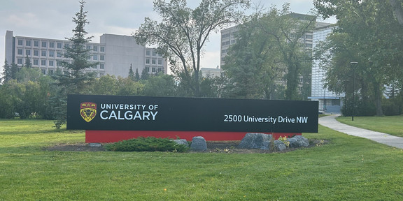 Foto: Campus der Universität Calgary. Schild mit der Aufschrift "University of Calgary" und der Adresse "2500 University Drive NW". Im Hintergrund sind mehrere Gebäude und Bäume sichtbar.
